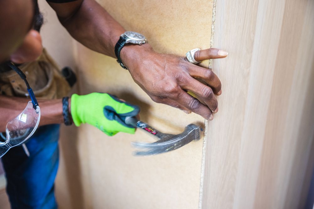 A carpenter's hands setting a trim board with a hammer and work gloves.